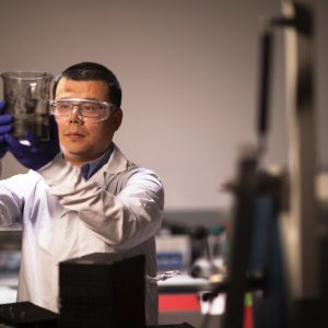 A man wearing a white lab coat and safety goggles holds up a beaker
