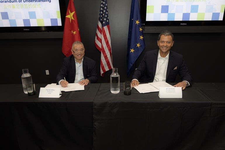 Two men sit at a table in front of flags of China, the EU, and the U.S.