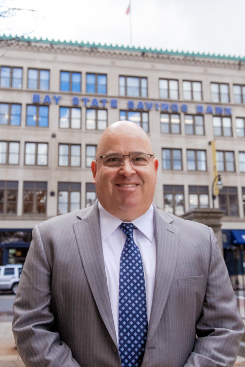 A man with glasses wears a grey pinstriped suit jacket, white button down, and blue patterned tie while standing in front of a tall grey building