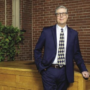 A man with grey hair wears grey glasses, a dark blue suit, white button down, and patterned tie while leaning on a wooden table top inside a brick building.