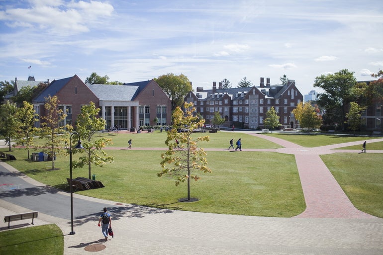 Outside a college campus with brick buildings and plots of green grass in front of them with students walking down the walkways in between the buildings