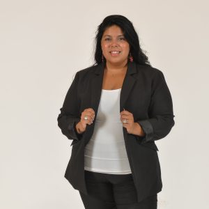A woman with long dark hair wears a black suit jacket and pants and a white top.