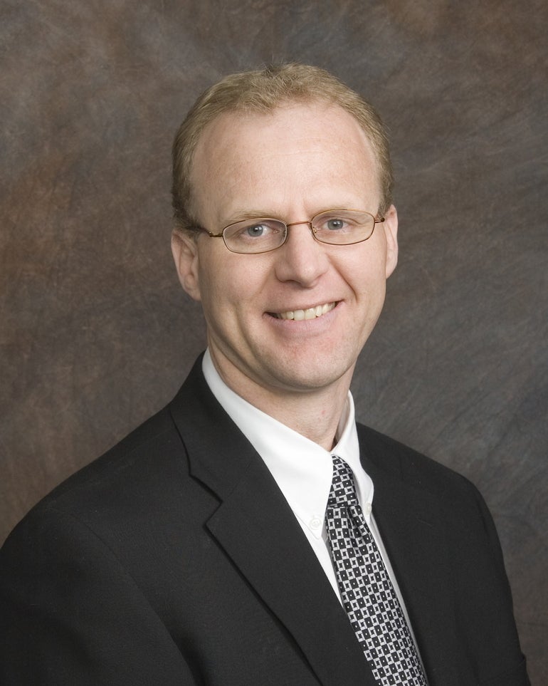 A man with short blonde hair wears thin wire-framed glasses, a black suit jacket, white button down, and black and white patterned tie.