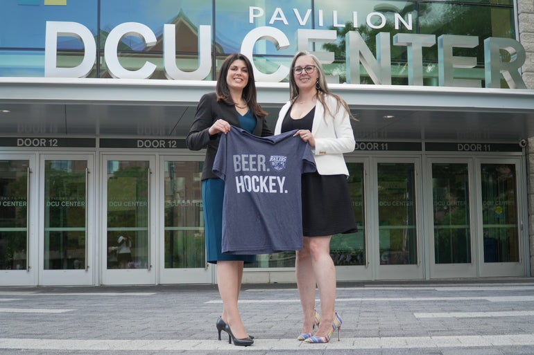 Two women in heels stand holding a t-shirt that says 