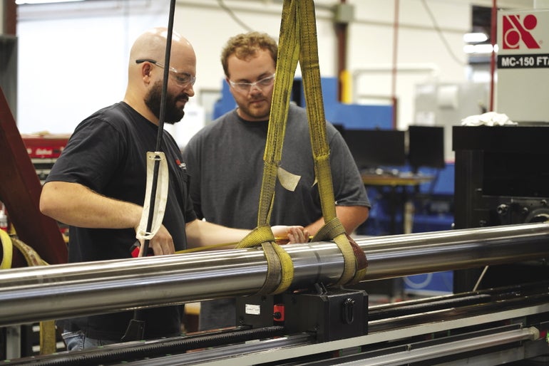 Two men wearing protective glasses stand behind a large metal pole held sideways by a yellow tether.