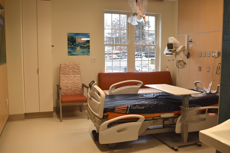 A hospital bed in a room with a red couch behind it and a high-back chair beside the couch with a window behind them looking out into a parking lot