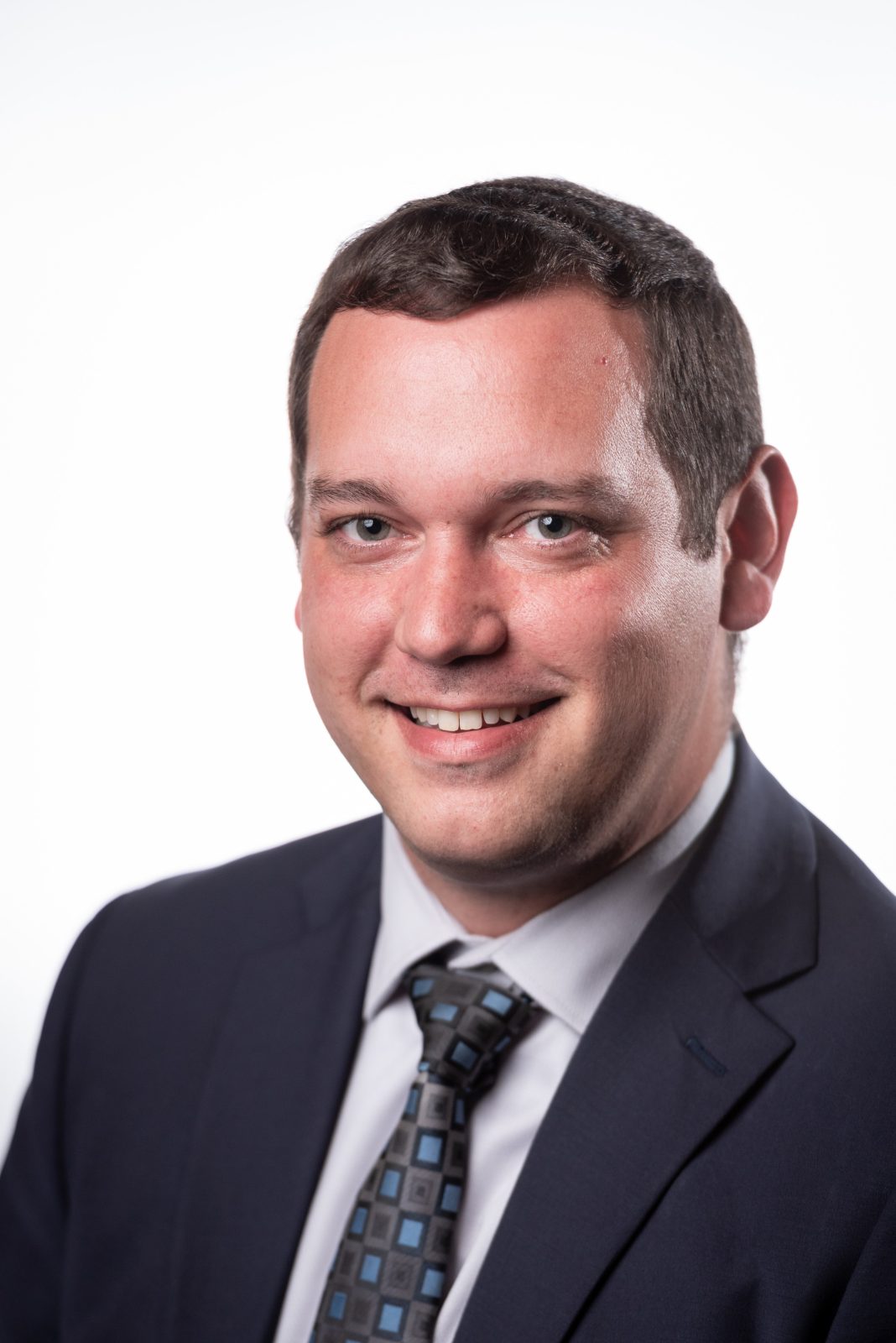 A man with short dark brown hair wears a dark navy suit, white button down, and grey and blue tie.