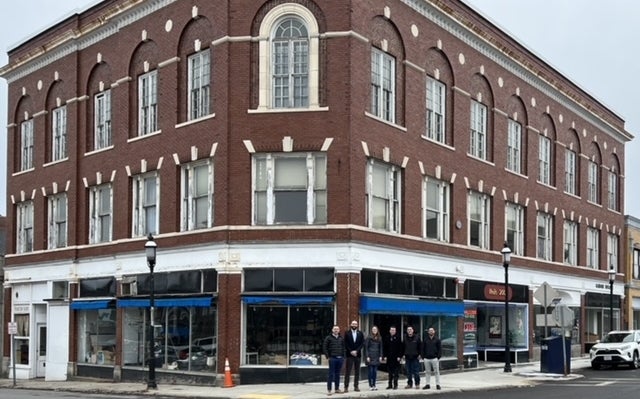 Six people stand in front of a brick building with blue awnings