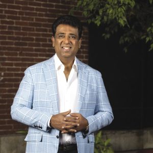 A man with short black hair wears a light blue plaid jacket, and a white button down while standing with his hands together in front of a brick building.