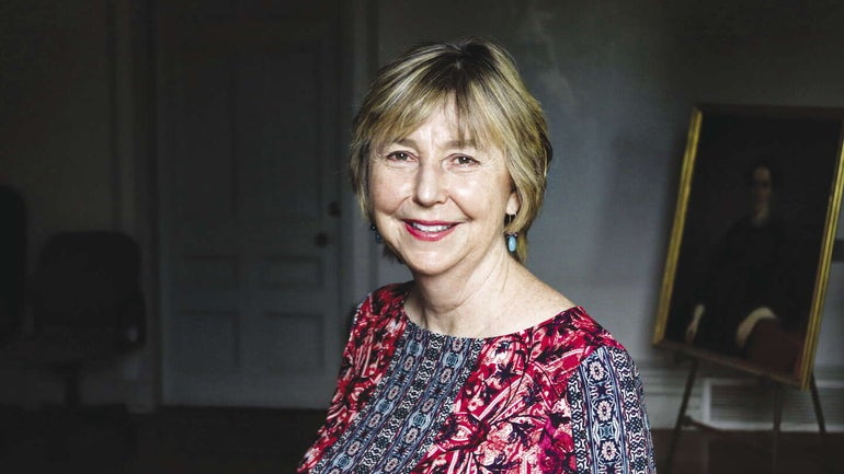 A woman with short blonde hair wears a red, black, blue, and white patterned top inside a dimly-lit room with a painting on a stand behind her.