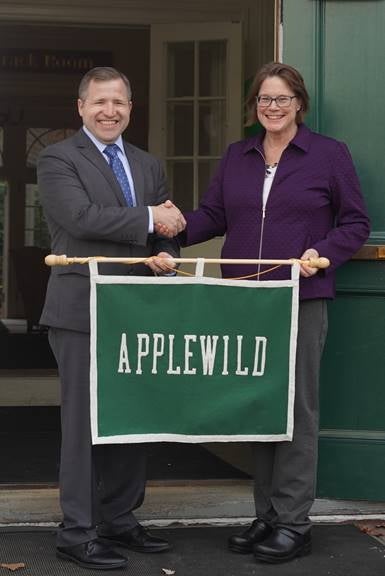 A man in a dark grey suit, light blue button down, and dark blue tie shakes hands with a woman in a purple jacket and dark grey pants while they both hold a green sign that reads 