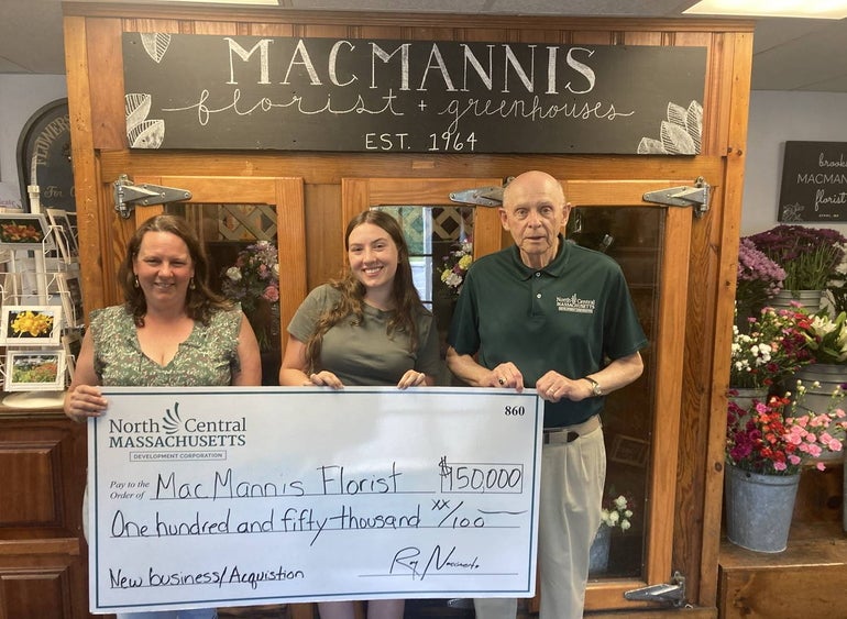 Two women an a man stand holding a giant check in front of a large wooden display cabinet with a sign on it reading 