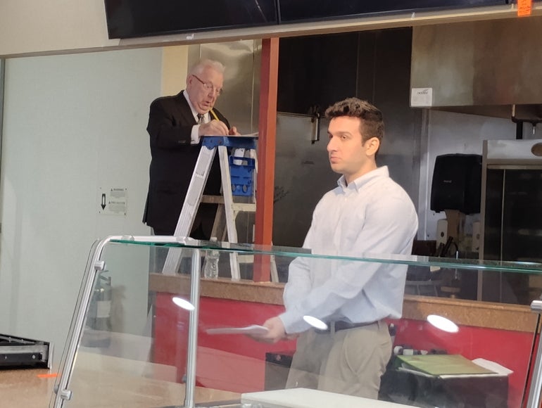 A man in dress clothes stands behind a sneeze guard, as a man in a suit stands on a ladder behind him.