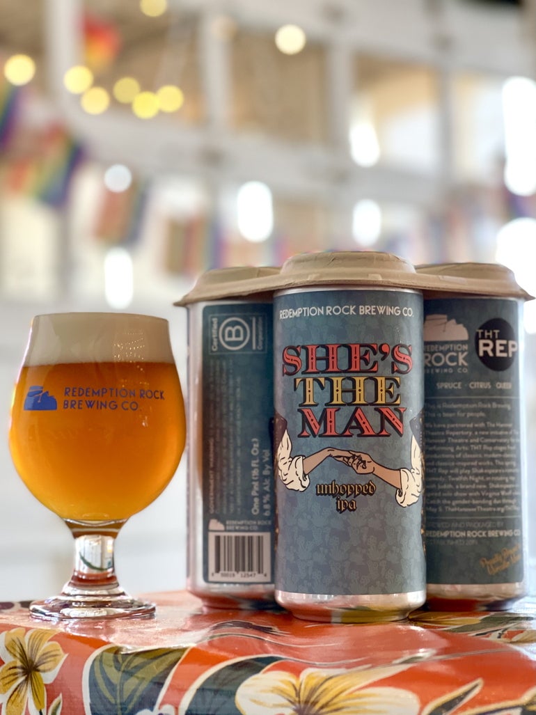 A glass of beer sits to the right of a group of three blue beer cans, the center can showing a cartoon image of hands being held underneath the beer's name 