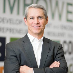 Man wearing a gray suit and white shirt without a tie has his arms folded and smiles at the camera.