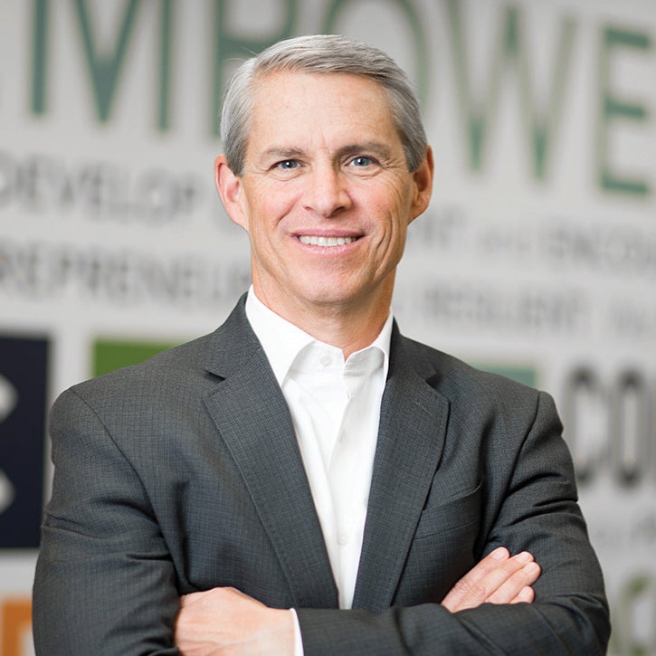 Man wearing a gray suit and white shirt without a tie has his arms folded and smiles at the camera.