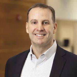 A man with short light brown hair wears a black blazer and da white checkered button down.