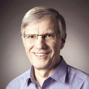 A  man with short grey hair wears glasses and a light purple button down.