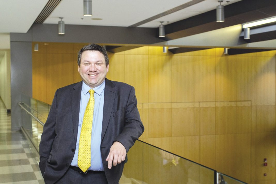 A man with short dark brown hair wears a dark grey suit jacket and pants, a light blue button down, and a yellow tie while leaning on a glass railing.