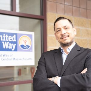 A man in a suit stands with his arms crossed in front of a window sign for the United Way