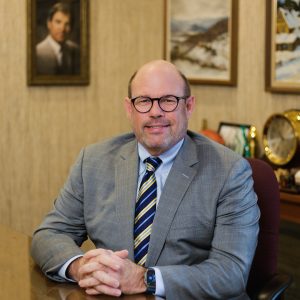 A man with glasses wearing a a gray suit sits at a table