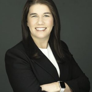 A woman with dark hair wears a black blazer, a white top, and an Apple watch.
