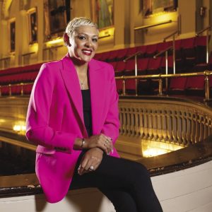 A woman with short blonde hair wears a neon pink suit jacket, black pants, and a black top while sitting in an auditorium.
