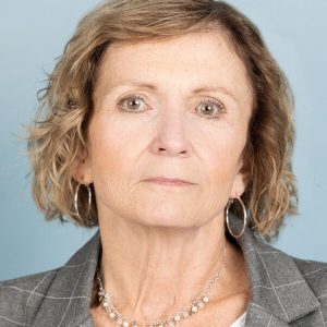 A woman with short, light hair is wearing a white shirt and gray jacket.