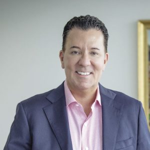 A man with short dark hair wears a dark blue suit jacket and pink and white checkered button down.