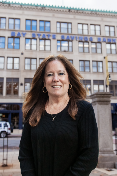 A woman with long brown hair wears a black blouse and gold hoops while standing in front of a building with 