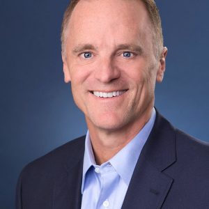 A man wearing a blue suit and blue shirt smiles at the camera.