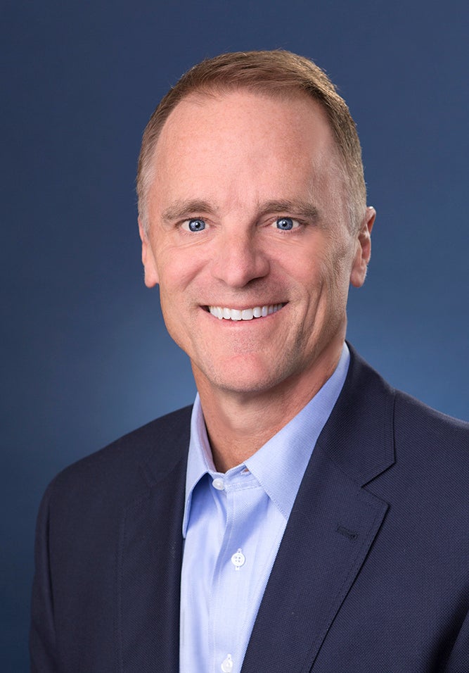 A man wearing a blue suit and blue shirt smiles at the camera.