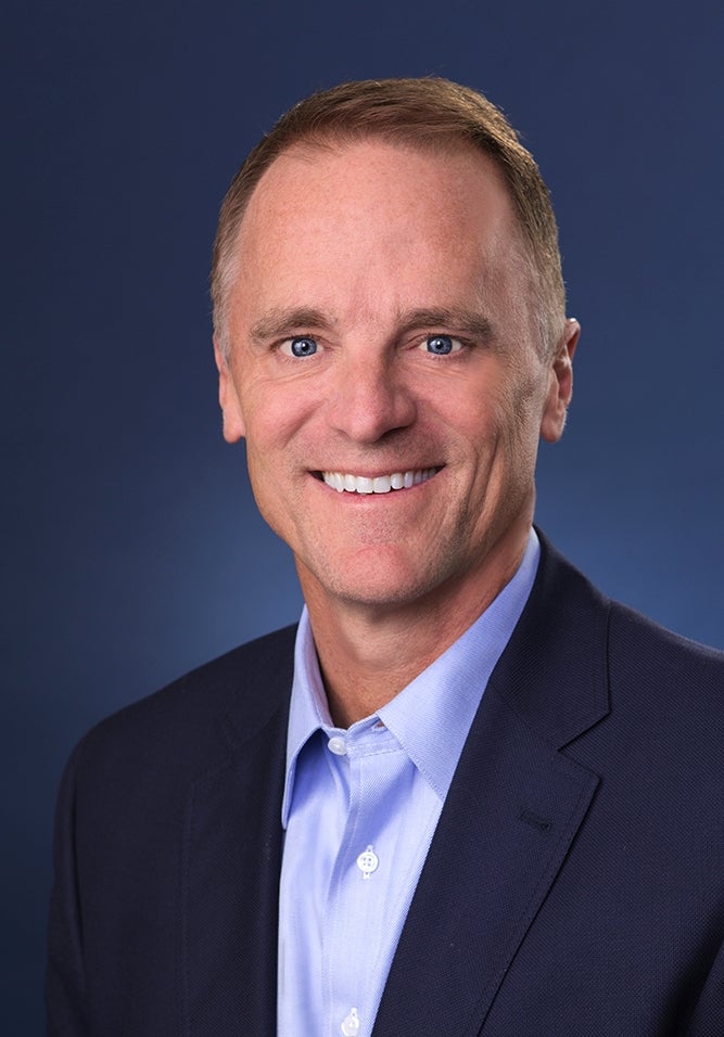 A man wearing a blue suit and blue shirt smiles at the camera.