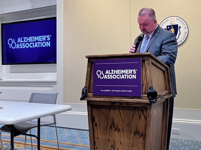 A man gets emotional while holding a microphone and standing behind a podium with the words 