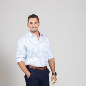 Man in dress clothes points at camera with both hands while smiling