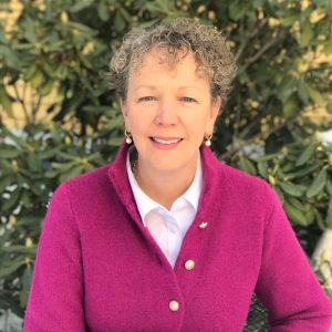 A woman with short, grey, curly hair wears a pink cardigan.