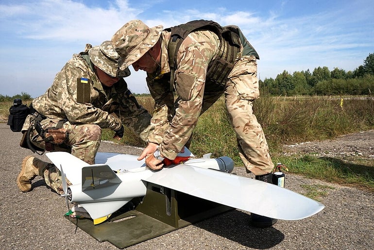 Two solders in camouflage fatigues  work on a small winged drone.