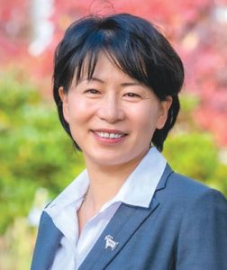 Headshot of a woman with short black hair wearing a blue suit jacket an white button down while standing in front of colorful leaves