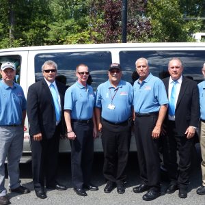 Seven men stand in line in front of a white van outside. The two on both ends of the line as well as the three in the middle wear light blue polos and the other two wear black suits, white button downs, and light blue ties.