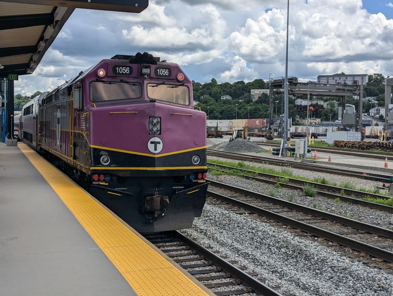 A purple train arriving at a passenger station with a railyard in the background