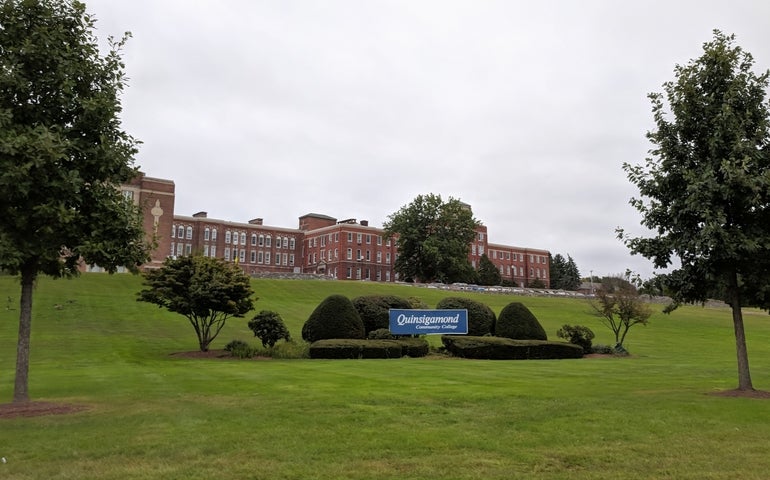 A brick university building is set back behind a large green lawn with trees and shrubs and a blue sign.