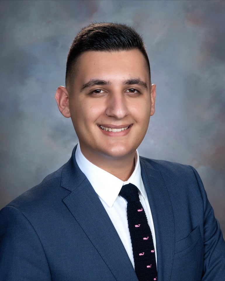 A man with short brown hair wears a dark blue suit jacket, a white button down, and a black tie with small pink whales on it.
