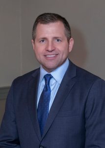 A man wearing a dark navy suit, light blue shirt, and blue tie smiles for the camera.