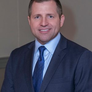 A man wearing a dark navy suit, light blue shirt, and blue tie smiles for the camera.