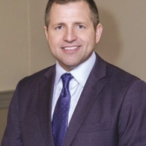 A man with short hair wears a dark blue suit jacket, white button down, and blue tie.