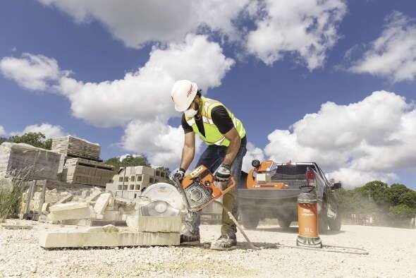 A man in construction gear holds an electric saw to a piece of concrete in front of a blue sky with clouds.