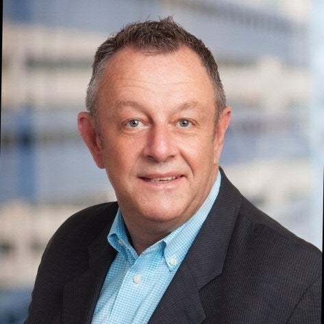 A headshot of a man in a dark grey suit jacket and light blue gingham button down