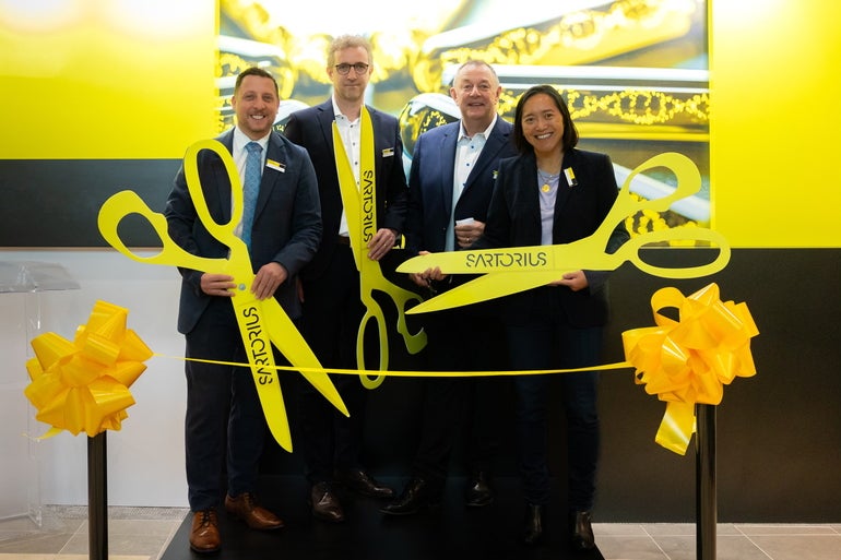 Four people in business attire hold oversized yellow scissors at a ribbon-cutting.