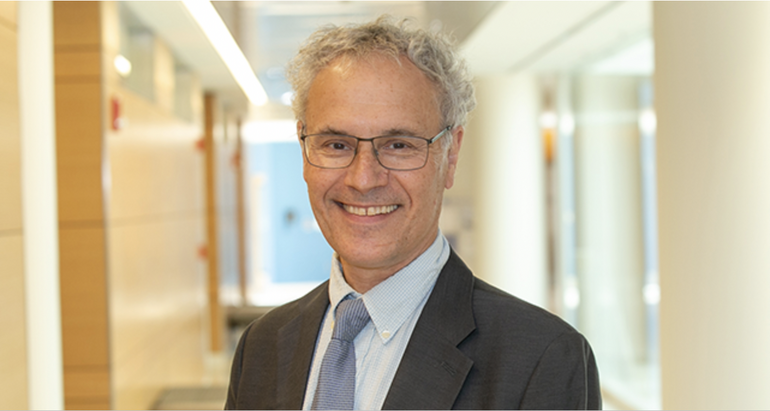 A man with curly grey hair wears glasses, a dark grey suit jacket, light blue button up, and blue striped tie while standing in a hallway.