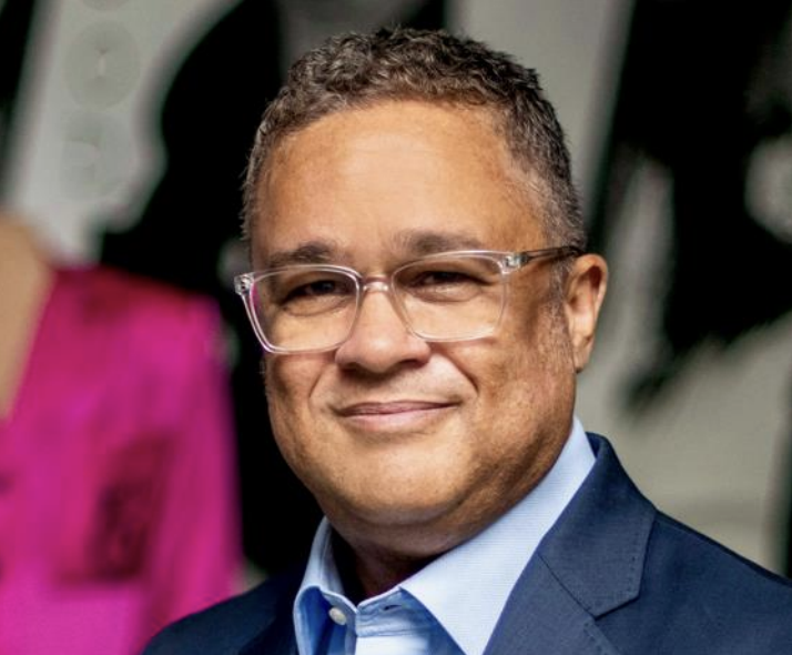 A man with short brown and grey hair wears clear glasses, a dark blue suit jacket, and light blue button down.
